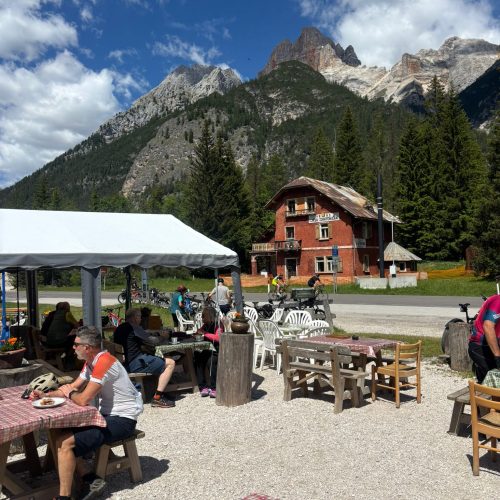 Alta Via 1 hikers relax at an outdoor cafe with Dolomites mountains in the background.